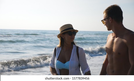 Happy Couple Of Lovers Walking At Seashore During Recreation On Resort. Man Hugs Woman Strolling Along Beach Enjoying Summer Vacation. Honeymoon Of Young Pair. Concept Of Love And Happiness. Slow Mo