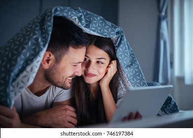 A Happy Couple Are Lying In Bed Under Covers While Watching A Romantic Film On The Tablet.