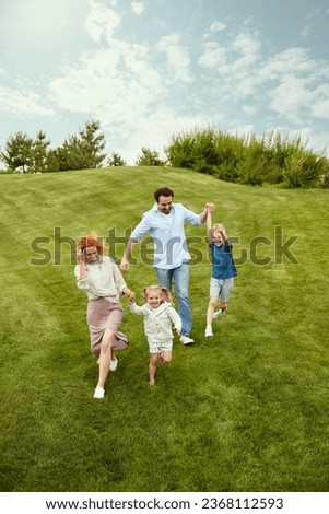 Happy couple, man and woman going for a walk in park with their little children, boy and girl, running, playing on grass. Concept of family, childhood and parenthood, fun, weekends, love, ad
