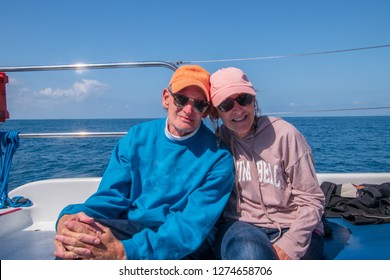 Happy Couple Of Older Caucasian Baby Boomers Sitting On A Boat With The Blue Water In The Background. Both Wearing Sweatshirts, Baseball Caps And Sunglasses