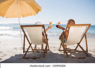 Happy couple on the beach drinking cocktails while relaxing on their deck chairs