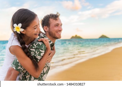 Happy Couple On Beach Vacation Travel Holiday In Hawaii. Asian Girl Laughing On Back Of Boyfriend Piggybacking Girlfriend In Lanikai, Oahu Island, Hawaii USA Destination.