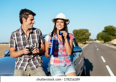 Happy Couple On Car Roadtrip Tourism. Man And Woman On Road Travel Vacation Taking A Break For Drinking And Taking Photos.