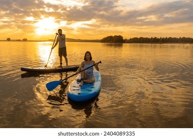 Happy couple paddle boarding at lake during sunset together with pug dog. Concept of active family tourism and supping with pets. Brave Dog Standing on SUP Board and enjoy lifestyle on summer vacation - Powered by Shutterstock