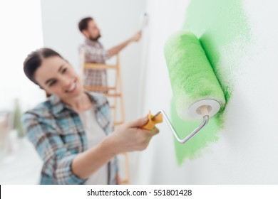 Happy Couple Painting Walls In Their New House: The Man Is Standing On A Ladder And The Woman Is Using A Paint Roller And Applying Bright Green Paint