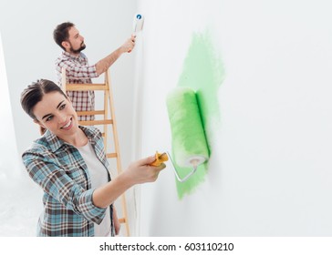 Happy Couple Painting Walls In Their New House: The Man Is Standing On A Ladder And The Woman Is Using A Paint Roller And Applying Bright Green Paint