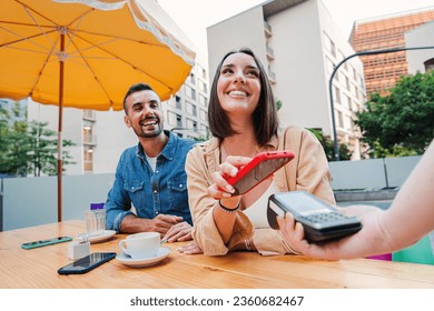 Happy couple paying the bill of the coffee shop using a cellphone contactless payment application. Young adult customer woman doing a purchase on a restaurant with a contactless smartphone banking app - Powered by Shutterstock