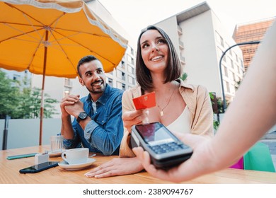 Happy couple paying the bill of the coffee shop using a contactless creditcard payment. Young adult customer woman doing a purchase on a restaurant to the cashier with a debit card. People spending - Powered by Shutterstock