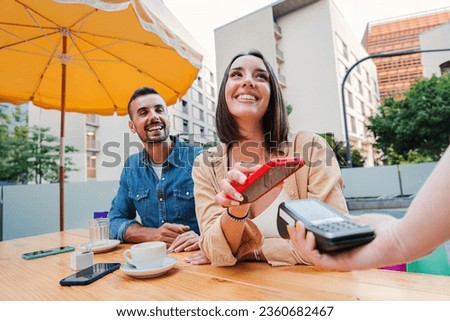 Happy couple paying the bill of the coffee shop using a cellphone contactless payment application. Young adult customer woman doing a purchase on a restaurant with a contactless smartphone banking app