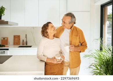 Happy couple of pensioners drinking coffee at kitchen, cheerful elderly husband and wife in casual outfits standing by kitchen table with mugs, having conversation and smiling, copy space - Powered by Shutterstock