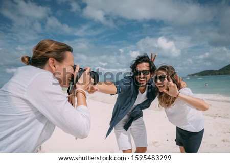 Happy couple and photographer on a tropical beach. Photoshoot with a professional photographer on a background of white sand, sea and cloudy sky. Newlyweds are photographed on a honeymoon at a resort