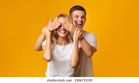 Happy Couple Playing Hide And Seek. Young Man Standing Behind His Girlfriend And Closing Her Eyes, Yellow Background With Copy Space