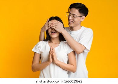 Happy Couple Playing Hide And Seek. Asian Man Standing Behind His Wife And Closing Her Eyes, Yellow Background