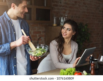 Happy couple preparing dinner with recipe, using tablet in their loft kitchen at home. Preparing vegetable salad.