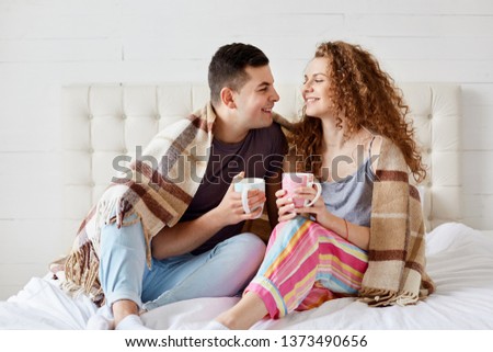 Happy couple relaxing in bed with hot beverage, holds cups full of tea or coffee, sit under warm blanket, dresses casually, romantic young family, spend spare time together at home. Relation concept.
