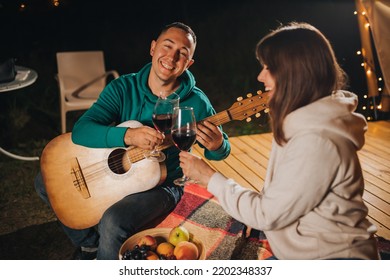 Happy Couple Relaxing In Glamping On Autumn Evening, Drinking Wine And Playing Guitar Near Cozy Bonfire. Luxury Camping Tent For Outdoor Recreation And Recreation. Lifestyle Concept