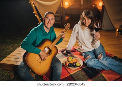 Happy Couple Relaxing In Glamping On Autumn Evening, Drinking Wine And Playing Guitar Near Cozy Bonfire. Luxury Camping Tent For Outdoor Recreation And Recreation. Lifestyle Concept