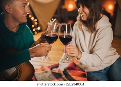 Happy Couple Relaxing In Glamping On Autumn Evening, Drinking Wine And Playing Guitar Near Cozy Bonfire. Luxury Camping Tent For Outdoor Recreation And Recreation. Lifestyle Concept