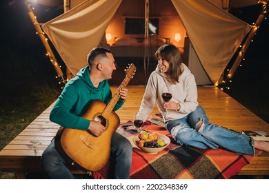 Happy Couple Relaxing In Glamping On Autumn Evening, Drinking Wine And Playing Guitar Near Cozy Bonfire. Luxury Camping Tent For Outdoor Recreation And Recreation. Lifestyle Concept