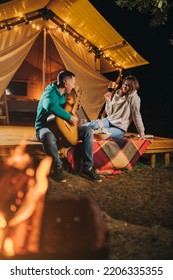 Happy Couple Relaxing In Glamping On Autumn Evening, Drinking Wine And Playing Guitar Near Cozy Bonfire. Luxury Camping Tent For Outdoor Recreation And Recreation. Lifestyle Concept