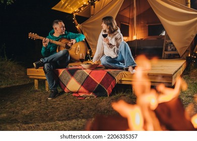 Happy Couple Relaxing In Glamping On Autumn Evening, Drinking Wine And Playing Guitar Near Cozy Bonfire. Luxury Camping Tent For Outdoor Recreation And Recreation. Lifestyle Concept