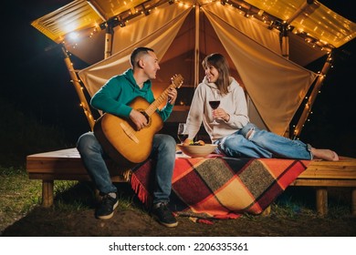 Happy Couple Relaxing In Glamping On Autumn Evening, Drinking Wine And Playing Guitar Near Cozy Bonfire. Luxury Camping Tent For Outdoor Recreation And Recreation. Lifestyle Concept