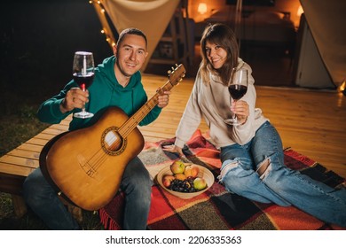 Happy Couple Relaxing In Glamping On Autumn Evening, Drinking Wine And Playing Guitar Near Cozy Bonfire. Luxury Camping Tent For Outdoor Recreation And Recreation. Lifestyle Concept