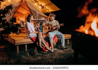 Happy couple relaxing in glamping on autumn evening, drinking wine and playing guitar near cozy bonfire. Luxury camping tent for outdoor recreation and recreation. Lifestyle concept - Powered by Shutterstock