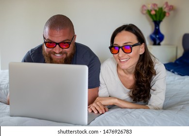 Happy Couple Relaxing At Home With Lap Top, Looking At The Screen With Orange Blue Light Blocking Glasses