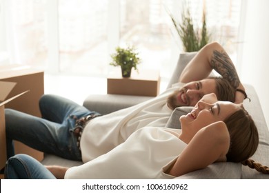 Happy couple relaxing on sofa having fun on moving day, excited young homeowners enjoying relocation in into new home, positive laughing man and woman sitting on couch with packed cardboard boxes