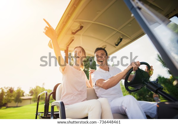 Happy couple riding a golf cart. A man in a\
white suit is sitting at the wheel and driving a golf car, a woman\
in a light suit is sitting beside\
him