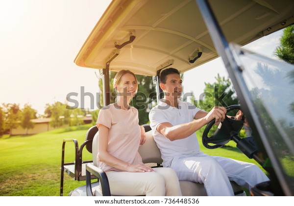 Happy couple riding a\
white golf cart on the golf course. A man in a white suit is\
sitting at the wheel, a woman in light clothes is sitting beside\
her and smiling