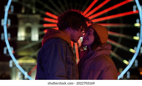 Happy Couple Rubbing Noses Against Ferris Wheel Outdoor. Cheerful Man And Woman Smiling On Urban Street. Affectionate Pair Having Romantic Date In City Center At Night.