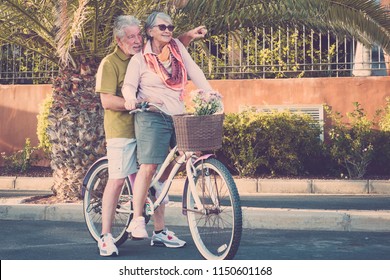 Happy Couple Of Senior Adult People Caucasian Go And Have Fun Together On The Same Vintage Bike In Outdoor Lsieure Activity. Tropical Place And Vacation Lifestyle For Beautiful Retired Man And Woman