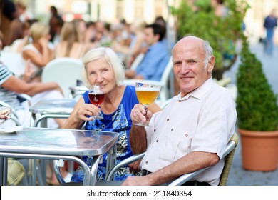 Happy couple of seniors, a man and his wife, are enjoying glass refreshing drink sitting at the open air terrace of cozy cafe in typical european city on a warm sunny day - active retirement concept