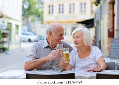 Happy couple of seniors, a man and his wife, are enjoying glass refreshing drink sitting at the open air terrace of cozy cafe in typical european city on a warm sunny day - active retirement concept
