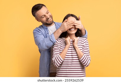 A happy couple shares a lighthearted moment as the man playfully covers the woman's eyes with his hands, both smiling broadly. They stand together against a bright yellow backdrop, radiating love. - Powered by Shutterstock