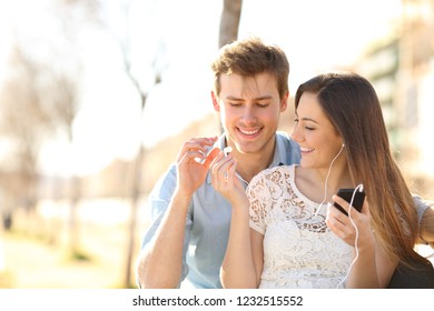 Happy Couple Sharing Earbuds To Listen To Music From A Smart Phone Sitting On A Bench In A Park