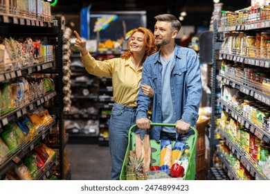 Happy couple with shopping cart walking at supermarket and buying food , woman pointing at shelf with goods, ask husband to buy something - Powered by Shutterstock