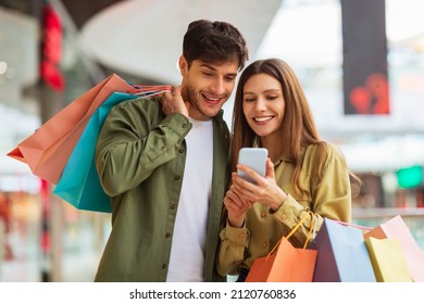 Happy Couple Shopping Using Mobile Phone Application Holding Colorful Shopper Bags And Buying Clothes In Modern Mall On Weekend. Customers Hold Smartphone Browsing Internet For Sales. Ecommerce