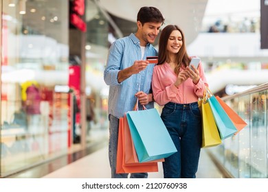 Happy Couple Shopping Using Smartphone And Credit Card Standing In Mall, Holding Colorful Shopper Bags. Buyers Using App Purchasing Clothes Online. Ecommerce Application Concept. Selective Focus