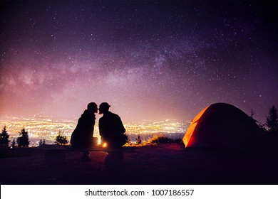 Happy Couple In Silhouette Kissing Near Campfire And Orange Tent. Night Sky With Milky Way Stars And City Lights At Background.