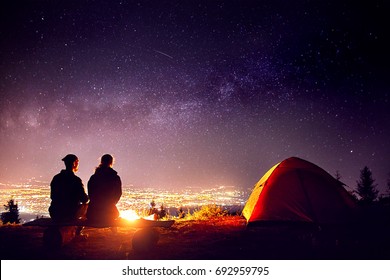 Happy Couple In Silhouette Sitting Near Campfire And Orange Tent. Night Sky With Milky Way Stars And City Lights At Background.