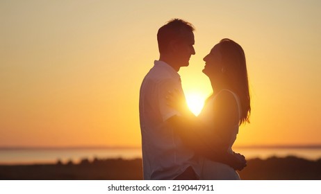 Happy Couple Silhouettes Stand Showing Pure Love To Each Other In Back Lit. Wife And Husband Enjoy Being Together And Stand On Meadow At Orange Back Sunset Side View