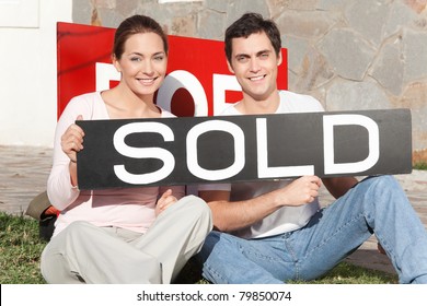 Happy Couple Sitting Against Their New House With Sold Sign