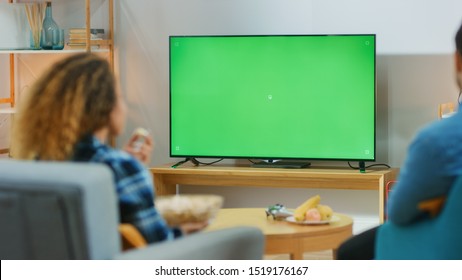 Happy Couple Sitting At Home In The Living Room Watching Green Chroma Key Screen TV, Relaxing On A Couch. Couple Room Watching Sports Match, News, Show Or A Movie.