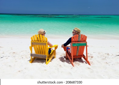 Happy Couple Sitting On Colorful Adirondack Chairs At Tropical Beach Enjoying Caribbean Vacation 