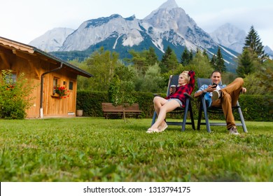 Happy Couple Sitting On Deck Chairs In Mountain Family, Travel, Tourism And People Concept. Vacation Relax Time In Nature With Sunlight.