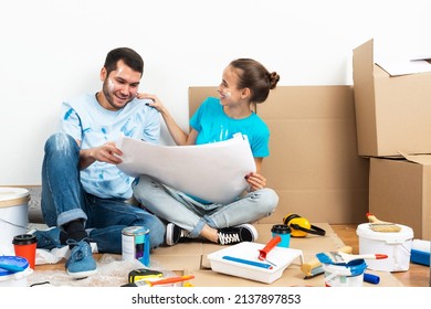 Happy Couple Sitting On Floor With Construction Blueprint. Home Remodeling And House Interior Redesign. Construction Tools And Materials Lying On Floor. Young Family Studies Renovation Project Of Flat