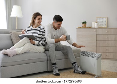 Happy Couple Sitting On Sofa Near Electric Heater At Home